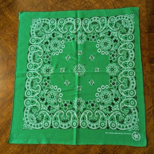 May include: Green bandana with a white and black paisley floral design. The square cloth features a repeating pattern of flowers and teardrop shapes. The bandana is made of 100% cotton.