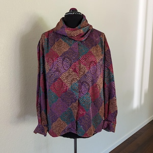 May include: A long-sleeved blouse with a cowl neck, displayed on a mannequin. The blouse features a patchwork design in shades of purple, brown, teal, and pink. The fabric has a textured, dotted pattern. The sleeves have buttoned cuffs.