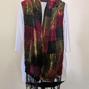 May include: A long rectangular scarf featuring a patchwork design of velvet-textured panels in deep red, gold, and black. The fabric displays various geometric patterns and linear motifs throughout, finished with long black fringe at both ends.