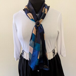 May include: A square scarf featuring a geometric pattern of navy blue, teal, and beige squares, secured with a dark brown four-petaled ring. The scarf is draped over a white three-quarter sleeve top and a black skirt with a white patterned waistband.