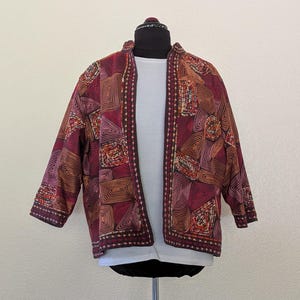 May include: An open-front jacket featuring a deep red and orange color palette with a geometric pattern of swirling lines and rectangular shapes. The garment has three-quarter length sleeves and a decorative stitched border along the front edges.