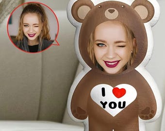 Op de afbeelding: Een bruin teddybeer-vormig kussen met het gezicht van een persoon in het midden. Het kussen heeft een wit hart met "I ❤️ YOU" erop gedrukt. Een kleinere afbeelding van de persoon staat in een rode cirkel linksboven.