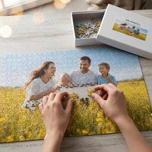 Op de afbeelding: Een puzzel met een familiefoto die in elkaar wordt gezet. De afbeelding toont een gezin in een veld met gele bloemen. Een witte doos met de tekst "APLANGGifts" en een foto van de familie staat in de buurt.