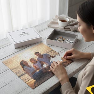 Op de afbeelding: Een witte doos met de tekst "APLANGGifts" staat naast een gedeeltelijk geassembleerde legpuzzel. De puzzel toont een familiefoto. Een persoon plaatst een puzzelstukje. Een kopje thee staat op de achtergrond.