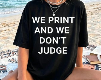 Op de afbeelding: Een zwart t-shirt met korte mouwen en de witte tekst WE PRINT AND WE DON'T JUDGE in het midden op de borst. Het shirt heeft een ronde hals en een relaxte pasvorm, gecombineerd met een gelaagde zilverkleurige ketting met een dunne hanger.