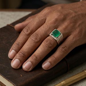 Può includere: Un anello d'argento con una grande gemma di smeraldo quadrata. L'anello è su una mano appoggiata su un taccuino di pelle marrone. Una penna stilografica d'oro è in primo piano.