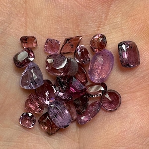 May include: A collection of faceted gemstones in shades of purple, pink, and brown. The gemstones vary in shape, including round, oval, and rectangular cuts. The gems are displayed in a hand, showcasing their color and clarity.