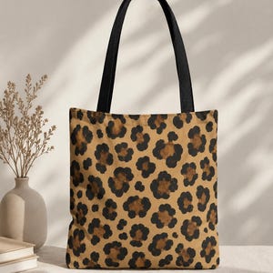 May include: A rectangular tote bag featuring a tan background with a repeating black and dark brown leopard print pattern. The bag includes two sturdy black fabric shoulder straps attached to the top edge, providing a functional design for carrying.