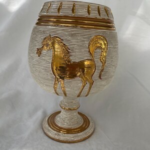 May include: A decorative, off-white goblet with a gold horse design. The goblet has a textured, horizontal striped pattern with gold accents around the rim and base. The horse is in a running pose.