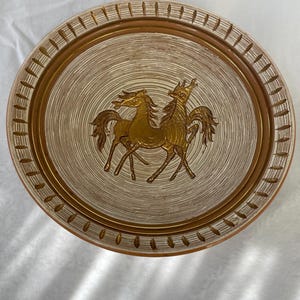 May include: An oval-shaped decorative tray with a gold horse design. The tray has a light beige center with concentric circle patterns and a brown border with vertical indentations. The horses are gold-colored and are the focal point of the tray.