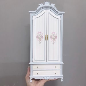 May include: A miniature white and light blue armoire with two drawers. The doors are decorated with pink floral designs and gold-colored handles. The armoire has a decorative top and base, and is held by a hand.