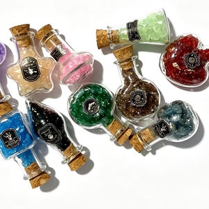Op de afbeelding: Een assortiment glazen flessen met kurken, gevuld met kleurrijke kristallen. De flessen hebben verschillende vormen, waaronder een hart, een ster en een vierkant. Elke fles heeft een etiket met een decoratief ontwerp.