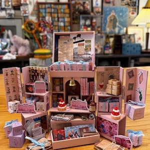 Op de afbeelding: Een miniatuur roze vitrinekast met meerdere compartimenten, gevuld met kleine cadeautjes, boeken en decoratieve items. De display bevat kleine dozen vastgebonden met blauwe linten, miniatuur desserts en vintage-achtige ephemera. Het woord "MENDLS" is zichtbaar.