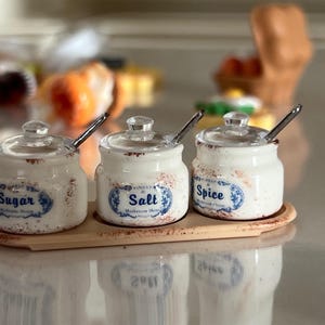 Op de afbeelding: Een set van drie kleine witte keramische potjes met doorzichtige glazen deksels en metalen lepels, geplaatst op een bijpassend ovaal dienblad. Elk potje heeft een blauw label met de tekst Sugar, Salt of Spice en een bruine spikkelafwerking.