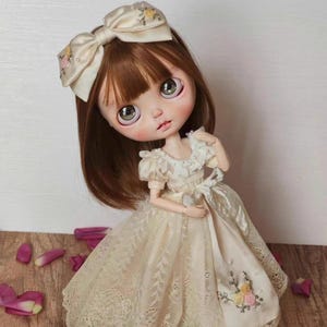 May include: A handmade doll with brown hair and large, expressive eyes. The doll wears a cream-colored dress with lace detailing and a matching bow in her hair. The dress features floral embroidery. The doll is a collectible item.