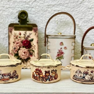 May include: A collection of miniature kitchenware with a vintage aesthetic. Includes small cream-colored pots, a clipboard with floral art, and decorative containers with handles. The pots have detailed illustrations.
