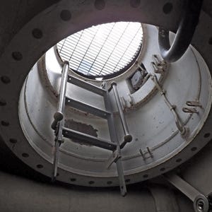 May include: Low-angle shot of a circular opening with a metal ladder. The opening is framed by a white, circular metal structure with bolt holes. A metal grate covers the opening, and the ladder is made of grey metal.