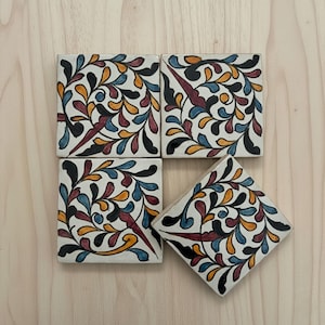May include: Four square ceramic tiles with a white background and a colorful floral design. The design includes black, blue, orange, and burgundy leaf-like shapes. The tiles are arranged on a light wood surface.