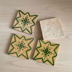 May include: Four square ceramic tiles. Three tiles feature a starburst design in green on a yellow background. The fourth tile is plain with a beige color. The tiles are arranged on a light wood surface.