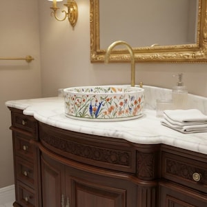 May include: A white ceramic basin with a floral pattern and a gold tap. The basin sits on a marble countertop above a dark wood vanity. A gold-framed mirror and sconces are in the background. The basin has a colourful floral design.
