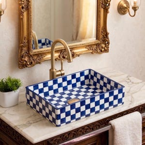 Può includere: Lavandino rettangolare in ceramica con motivo a scacchi blu e bianco. Il lavandino è posizionato su un piano in marmo con un rubinetto dorato. Uno specchio con cornice dorata è appeso sopra il lavandino e una piccola pianta in vaso è a sinistra.