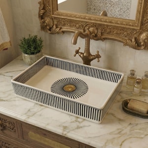 May include: A rectangular ceramic sink with a white base, black vertical stripes, and a central sunburst design. The sink is set on a marble surface, with a gold tap and mirror in the background.