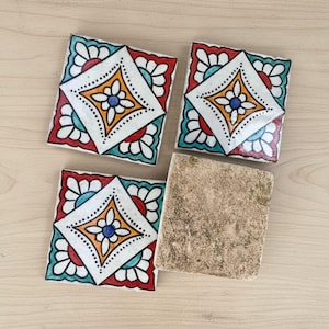 Puede incluir: Cuatro baldosas de cerámica cuadradas. Tres baldosas presentan un diseño en forma de diamante blanco con una flor central, rodeado de patrones florales naranjas, azules, rojos y blancos. Una baldosa tiene una superficie beige lisa y texturizada.