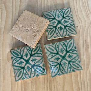 Puede incluir: Cuatro baldosas de cerámica cuadradas. Tres baldosas presentan un patrón floral verde sobre un fondo beige. Una baldosa es de color beige liso. Las baldosas están dispuestas sobre una superficie de madera marrón claro.