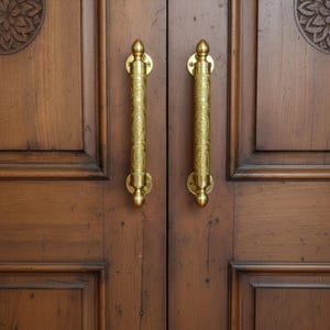 May include: Close-up of a pair of wooden doors with ornate brass handles. The doors are a rich brown color with visible wood grain. The handles are vertical, with detailed floral patterns.
