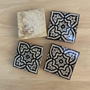 May include: Four square ceramic coasters. Three coasters feature a black and white floral pattern. The fourth coaster is a solid off-white color. The coasters are arranged on a light wood surface.