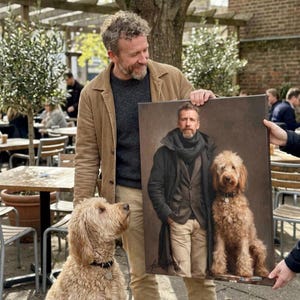 May include: A man holding a portrait of himself and a dog. The portrait is a painting of the man and a dog. The dog in the portrait is the same breed as the dog standing next to the man. The man is wearing a brown jacket and a gray sweater.