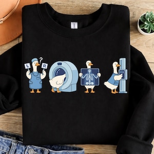 Könnte beinhalten: Schwarzes Sweatshirt mit medizinischem Thema und Cartoon-Enten. Eine Ente hält Schilder mit der Aufschrift „L“ und „R“, eine andere steht neben einem CT-Scanner, eine dritte zeigt ein Röntgenbild und die letzte bedient ein Röntgengerät.