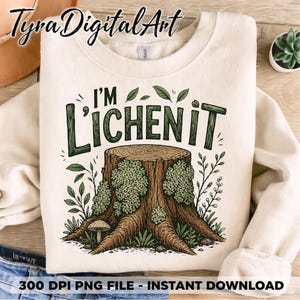Peut inclure: Sweat-shirt crème avec un motif vert d'une souche d'arbre et le texte "I'M LICHEN IT". Le motif comprend des feuilles vertes et un champignon. Le sweat-shirt est plié, avec un soupçon de jean bleu visible.
