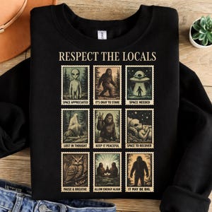 May include: Black sweatshirt with the text "RESPECT THE LOCALS" above nine postage stamp-style illustrations of aliens, Bigfoot, and UFOs. Each stamp has a unique caption, such as "SPACE APPRECIATED" and "IT MAY BE BIG."