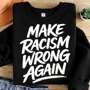 May include: Black sweatshirt with the white text "MAKE RACISM WRONG AGAIN" in a bold, handwritten style. The sweatshirt is displayed on a wooden surface with a pair of blue jeans and a small potted plant in the background.