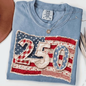 以下が含まれることがあります： 愛国的な旗のデザインが施された、水色のComfort Colors Tシャツ。旗には、赤、白、青の生地を使ったパッチワーク風の数字「250」が描かれています。シルバーのネックレスとペンダントがシャツの上に置かれています。