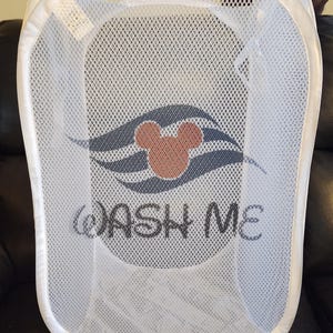 May include: White mesh laundry hamper with a white frame. The hamper features a design with a Mickey Mouse head in the center, above the words "WASH ME" in black script. The hamper is collapsible.