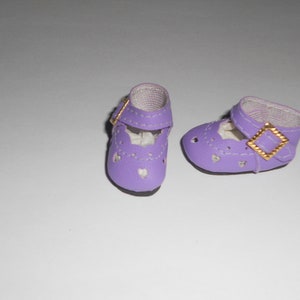 May include: A pair of lavender doll shoes with heart-shaped cutouts. The shoes have a strap with a gold-colored buckle. The soles are black. The shoes are designed to be worn by a doll.