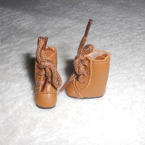 May include: A pair of brown leather ankle boots with laces tied in a bow. The boots are on a white fuzzy surface.