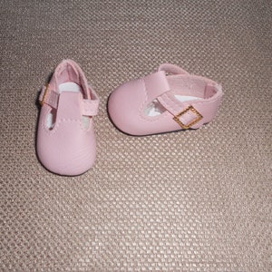 May include: A pair of pink baby shoes with a gold buckle. The shoes have a strap across the top and a closed toe.