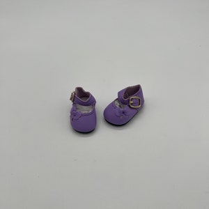 May include: A pair of purple doll shoes with gold buckles. The shoes have a strap across the top and a small bow on the toe.