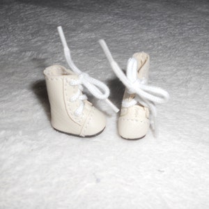 May include: A pair of white doll-sized boots with white laces. The boots are made of a soft, leather-like material.