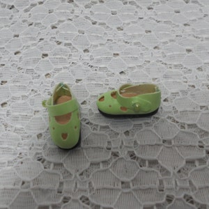 May include: A pair of green doll shoes with cutouts and a strap. The shoes have a black sole.