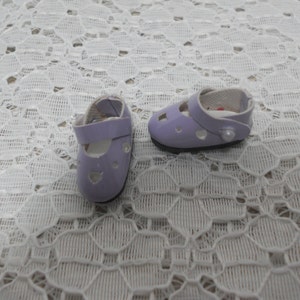 May include: A pair of lavender doll shoes with a strap and a small white button on each shoe.