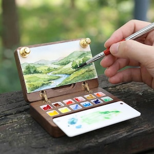 May include: A wooden watercolour paint set with a miniature landscape painting attached. The set includes a palette of various colours and a white mixing tray. A paintbrush is held over the painting.