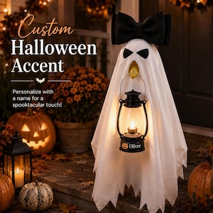May include: A white ghost decoration featuring a large black bow on top, black eye cutouts, and a gold-colored beak. The ghost holds a black lantern with a glowing light and the text "Oliver" printed next to a small orange pumpkin icon.
