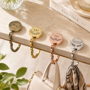 May include: Four circular metal bag hangers in antique brass, gold, rose gold, and silver finishes. Each hanger features a hinged, foldable hook and a flat top surface engraved with custom floral designs, initials, and the text Your text here.