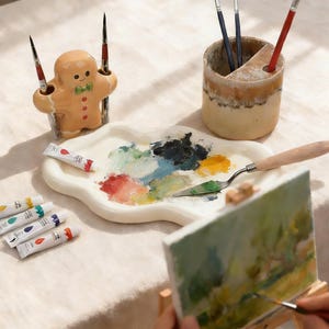 以下が含まれることがあります： 混合された絵の具の色、パレットナイフ、絵の具チューブが入ったセラミックアーティストのパレット。ジンジャーブレッドマン型のブラシホルダーと、筆付きのセラミックブラシホルダーも存在します。小さな絵画のあるイーゼルが前景にあります。