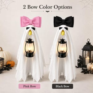 May include: Two ghost-shaped decorations featuring white fabric drapes, black eye patches, and yellow beaks. Each ghost holds a black lantern and wears a large bow on top, one in pink and one in black. The text 2 Bow Color Options is displayed above.