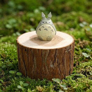 May include: A gray and white Totoro figurine with a light yellow belly, perched on a small, round wooden stump. The stump is surrounded by green moss. The figurine has pointed ears and distinctive markings.